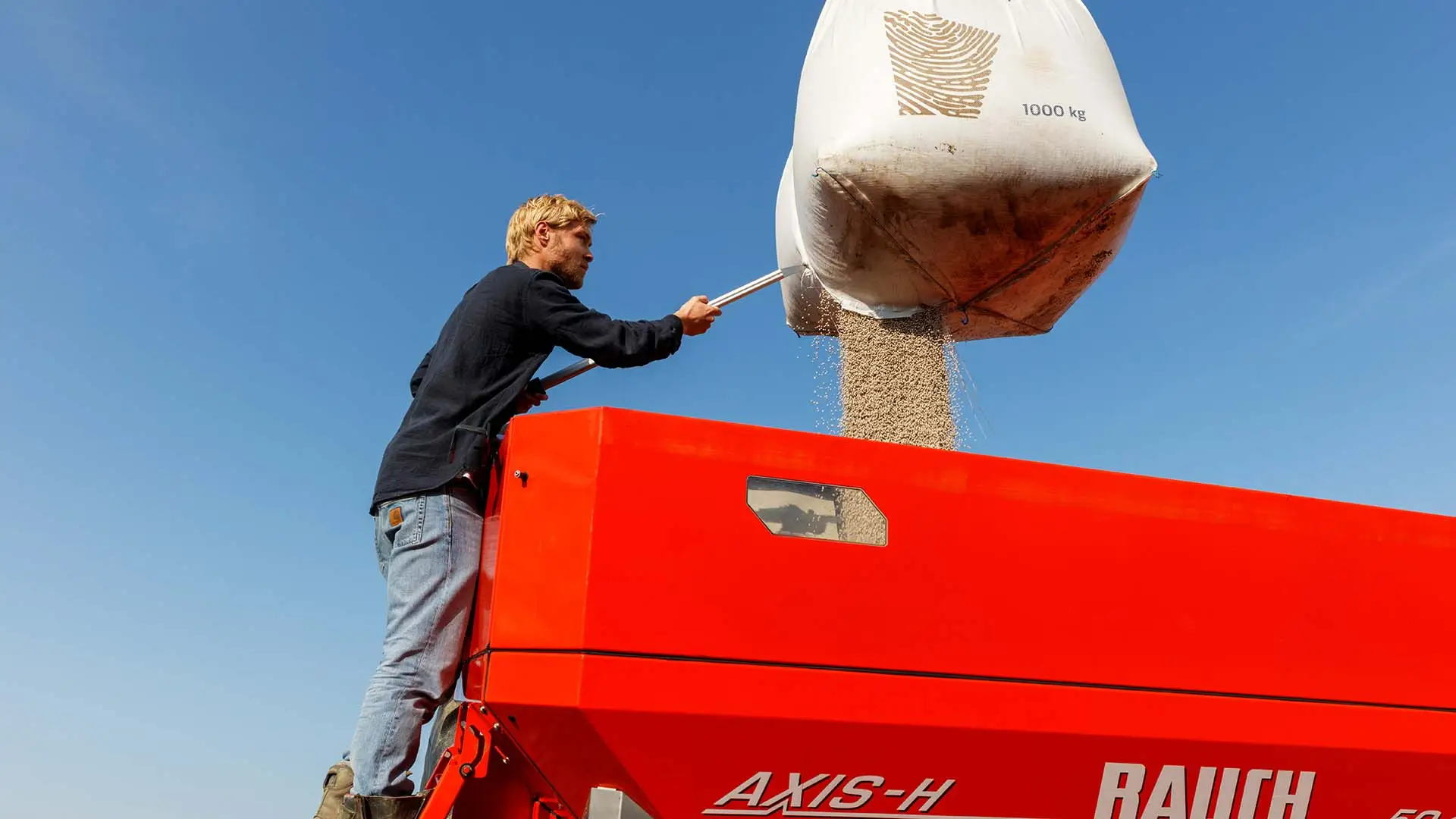 Landwirt öffnet von der Leiter aus einen Big Bag mit Dünger über einem roten Düngerstreuer.